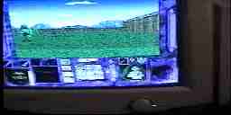 Close-up of a bulky 2003-era computer monitor displaying a computer game with a field near a building, a large HUD, and character face portraits.