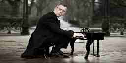 A man in a suit sitting at a tiny piano as he struggles to play it.