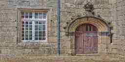 The exerior wall of an old stone bric building with an arch over the wooden door and a window nearby.